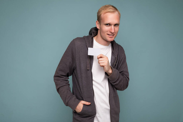 Knappe blonde man draagt grijze trui en wit t-shirt geïsoleerd over blauwe achtergrond muur met credit card kijken naar camera - Foto, afbeelding