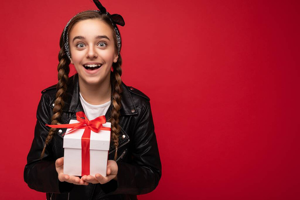 Photo of beautiful happy brunette girl isolated over red background wall wearing white casual t-shirt and stylish black leather jacket holding white gift box with red ribbon and looking at camera - Photo, Image