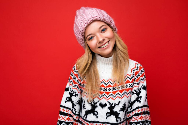 Photo de belle jeune femme blonde souriante et heureuse isolée sur un mur de fond rouge portant un pull d'hiver et un chapeau rose tendance regardant la caméra - Photo, image