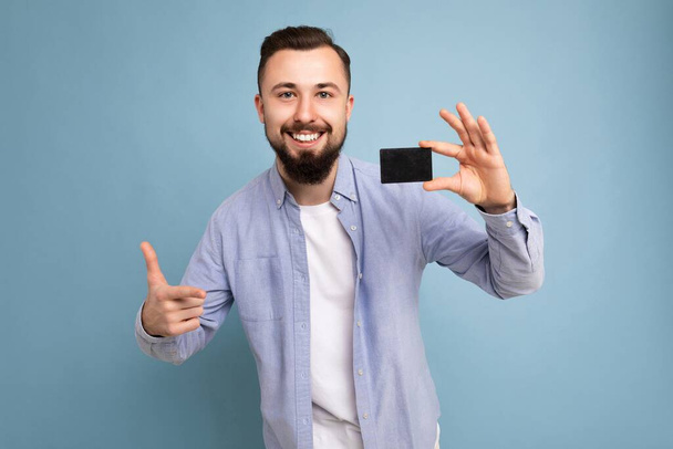 фото красивой улыбающейся брюнетки бородатого молодого человека в стильной синей рубашке и белой футболке, изолированной на синем фоне, держащей кредитную карту, смотрящей в камеру - Фото, изображение