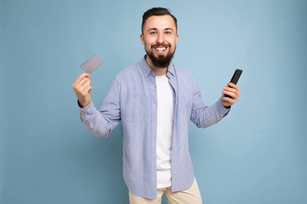 Fotografie dobře vypadající atraktivní úsměv brunet neoholený mladý muž na sobě ležérní modré košile a bílé tričko izolované přes modré pozadí zdi drží kreditní kartu a pomocí mobilního telefonu - Fotografie, Obrázek
