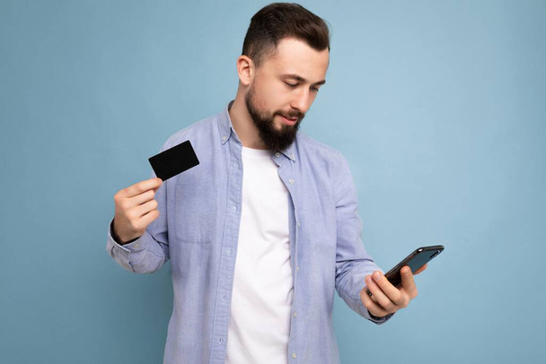 closeup Fénykép a jóképű vonzó mosolygós brunet borotválatlan fiatal férfi visel alkalmi kék ing és fehér póló elszigetelt kék háttér fal gazdaság hitelkártya és a mobiltelefon - Fotó, kép