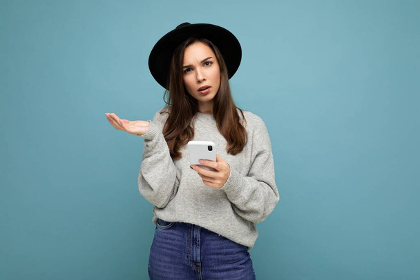 Красивая молодая спрашивает недовольная брюнетка женщина в черной шляпе и сером свитере держа смартфон глядя на камеру изолированы на заднем плане - Фото, изображение