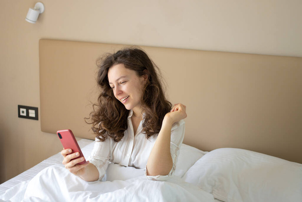 美しいブルネットの若い女性は、自宅で無線インターネット接続を使用して、朝の睡眠後に白いベッドに横たわってスマートフォンを見ている。友人とのチャット、ソーシャルメディアのチェック、メッセンジャー.  - 写真・画像