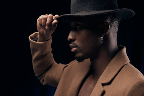 Portrait of a dark-skinned handsome man on a black background looking in front. Dressed in a brown jacket and a black hat holding his hand. - Foto, imagen