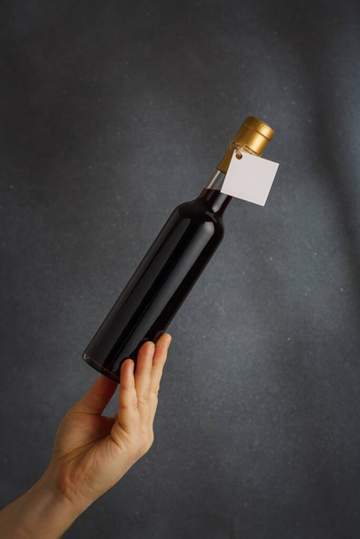 Male hand holding bottle of red wine, natural fruit alcohol liquor with empty label on grey background. Mock up, template for brand, vertical shot - Φωτογραφία, εικόνα