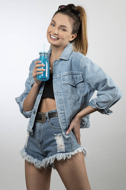 menina bonita sorrindo ela usa jaqueta e calções de ganga sorrindo com uma garrafa de bebida azul - Foto, Imagem