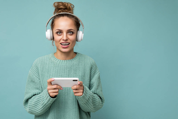 Closeup photo of beautiful positive happy young woman wearing blue sweater isolated over blue background wearing white bluetooth wireless earphones and listening to music and using mobile phone - Photo, Image