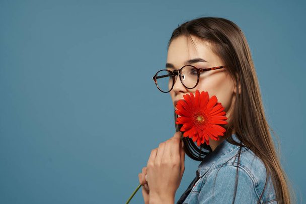 mooi brunette dragen bril rood bloem in de buurt gezicht Glamor blauw achtergrond - Foto, afbeelding