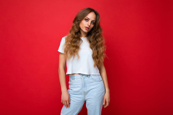 Young beautiful curly blonde woman with sexy expression, cheerful and happy face wearing casual white t-shirt isolated over red background with copy space - Photo, Image