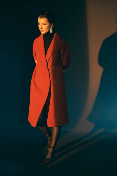 Pensive attractive girl in a red long coat on a dark background with folded arms behind looks forward - 写真・画像