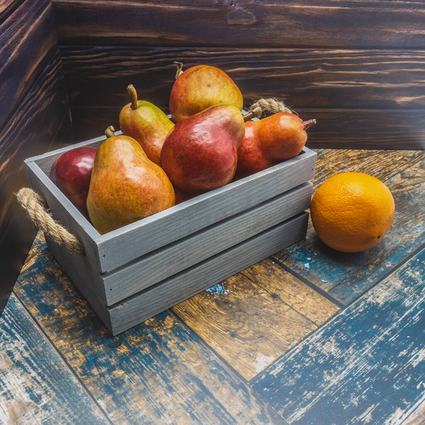 Una scatola di legno blu con manici di corda è piena di pere mature e mele. Nelle vicinanze, sulle tavole, si trova un'arancia. Primo piano - Foto, immagini