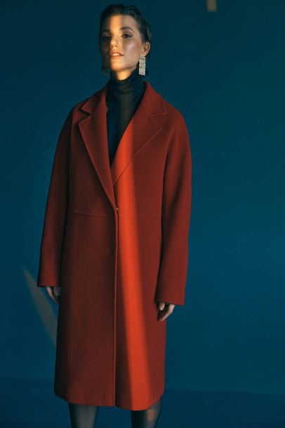 Girl in a red coat on a blue background. She stands with her arms evenly lowered and the sunlight falls on her - Foto, afbeelding