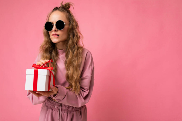 Shot van mooie positieve jonge blonde krullende vrouw geïsoleerd over roze achtergrond muur dragen roze sport kleding en zonnebril houden geschenkdoos en unboxing verrassing kijken naar de zijkant - Foto, afbeelding