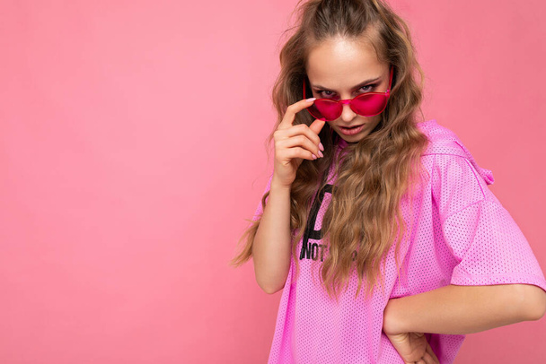 Okouzlující sexy vážné mladé blondýny kudrnaté ženy izolované na růžovém pozadí zdi na sobě ležérní růžové tričko a stylové barevné sluneční brýle při pohledu na kameru - Fotografie, Obrázek