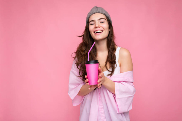 Mooie jonge positieve brunette vrouw dragen casual stijlvolle kleding geïsoleerd over kleurrijke achtergrond muur met papieren beker voor mockup drinken koffie kijken naar camera - Foto, afbeelding