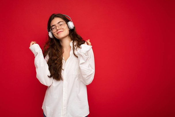 魅力的な幸せな笑顔若いブルネット白いシャツと眼鏡を身に着けている女性は、音楽やダンスを聞いて白い無線ブルートゥースイヤフォンを身に着けている赤い背景に隔離された - 写真・画像