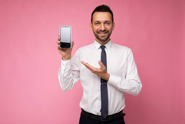 Fénykép jóképű mosolygós felnőtt férfi személy jóképű visel alkalmi ruhát áll elszigetelt a háttérben másolás tér gazdaság okostelefon mutatja, telefon a kezében üres képernyő kijelző - Fotó, kép