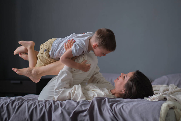 mom lying in bed have fun play with cute little son. Joyful young strong woman lifting on legs arms laughing little child , having fun together. - Photo, Image