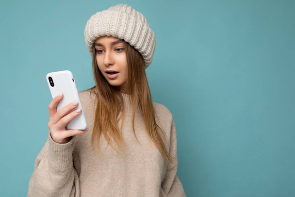 Photo of beautiful surprised young blonde woman wearing stylish beige warm sweater and knitted winter warm beige hat isolated on blue background with empty space holding in hand and using mobile phone - Foto, immagini