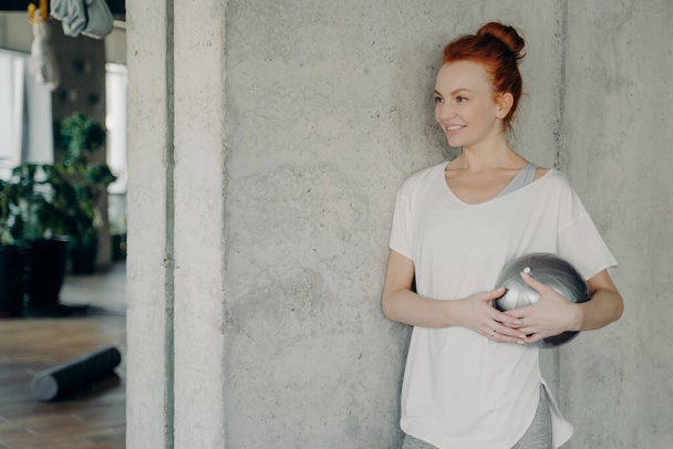 Positive ginger lady standing against gray wall with fitball in hand - Photo, Image