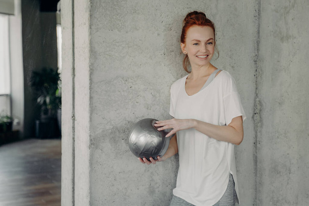 Red haired positive woman standing against gray wall with fitball in hand before pilates class - Photo, Image