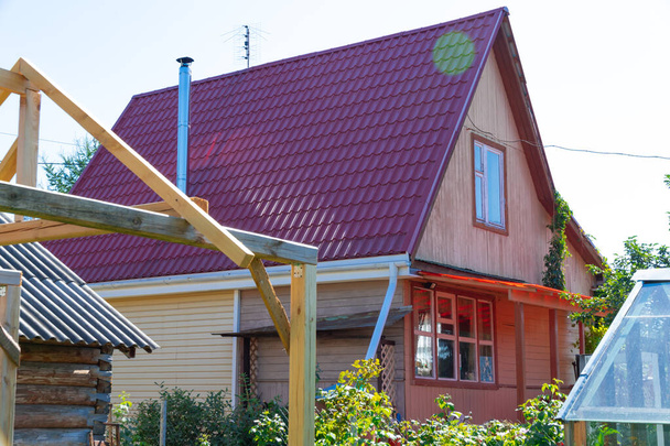 Dächer von Holzhäusern und Gewächshäusern im Dorf vor dem Hintergrund von blauem Himmel und grünem Laub. Heißer, sonniger Sommertag. Mittelfristiger Plan - Foto, Bild