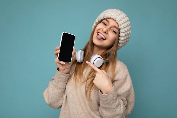 Фото сексуальної привабливої позитивної молодої темної блондинки в бежевому стильному светрі і в'язаному бежевому зимовому капелюсі ізольовані на синьому фоні в білому Bluetooth бездротові навушники і
 - Фото, зображення