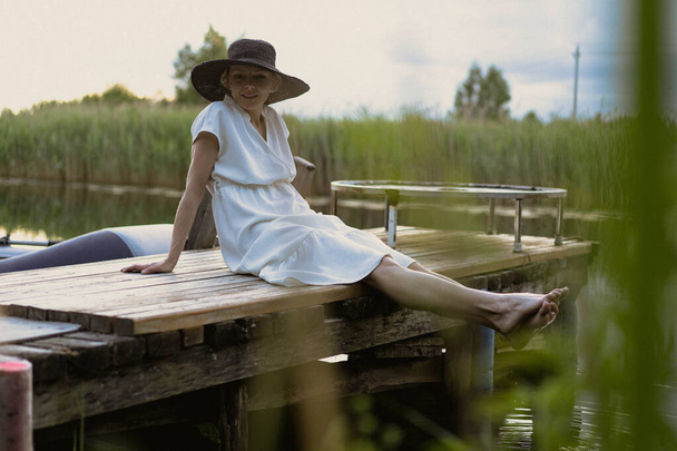 Młoda kobieta w sukience i kapeluszu w parku nad jeziorem na molo - Zdjęcie, obraz