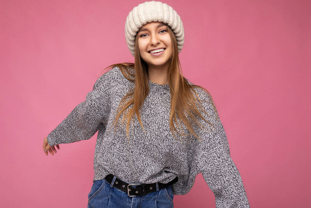Zdjęcie pięknej uśmiechniętej młodej ciemnej blondynki stojącej samotnie nad różową ścianą tła w szarym swetrze i zimowym beżowym kapeluszu patrzącej na aparat i bawiącej się - Zdjęcie, obraz