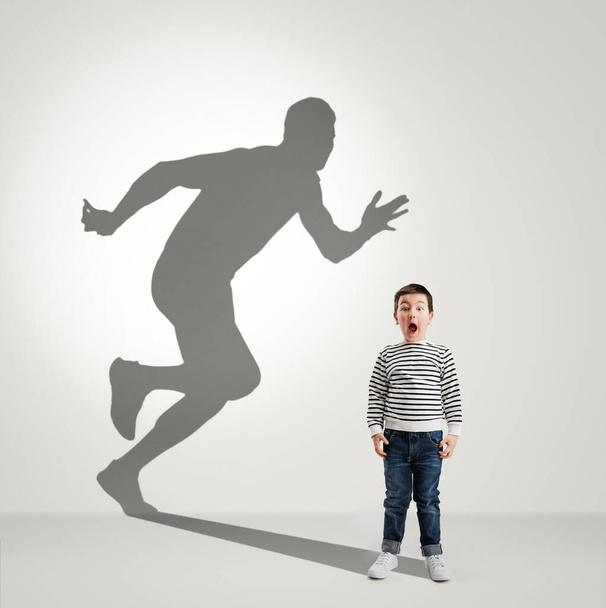 Childhood and dream about big and famous future. Conceptual image with boy and shadow of fit male runner on light gray background - Foto, immagini
