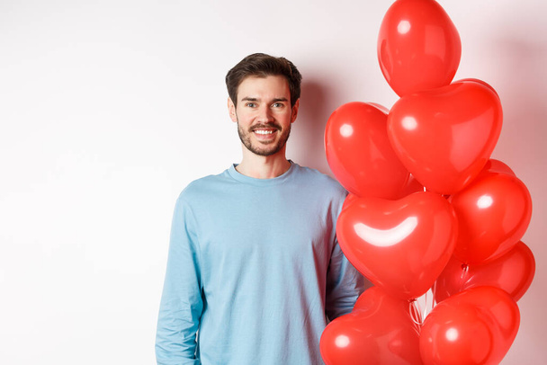 Smiling young man standing with heart balloons and looking happy, celebrating valentines day, bring romantic present to lover, standing over white background - Photo, Image