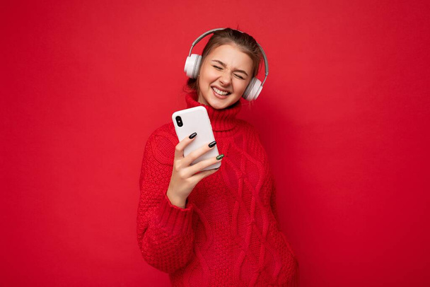 赤い背景の壁の上に隔離された赤いセーターを着て美しい幸せな若いブルネットの女性の写真を保持し、携帯電話を使用して、楽しんでいるBluetoothヘッドフォンを身に着けている - 写真・画像