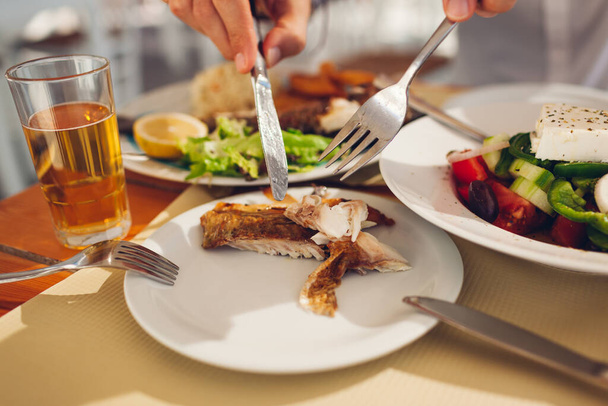 Człowiek kładzie kawałek ryby na talerzu z warzywami jedząc lunch w restauracji na świeżym powietrzu. Świeże owoce morza z sałatką grecką - Zdjęcie, obraz