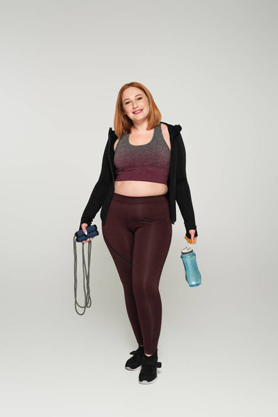 Cheerful plus size sportswoman holding skipping rope and sports bottle on grey background - Foto, immagini