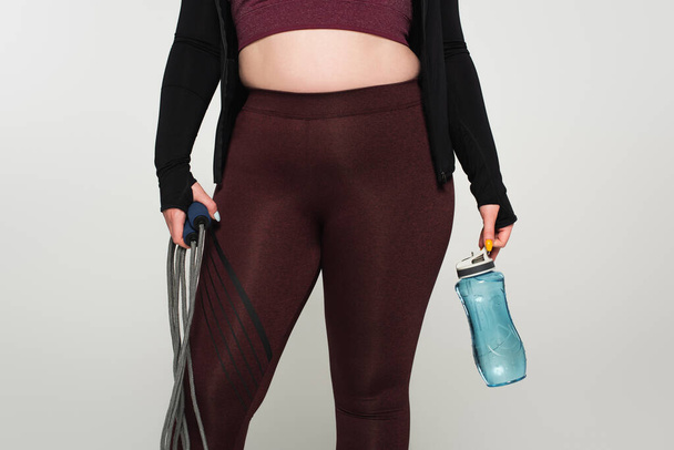 Cropped view of plus size sportswoman holding sports bottle and jump rope isolated on grey  - Photo, Image