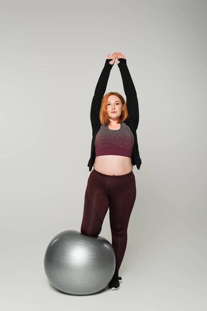 Redhead plus size sportswoman stretching near fitness ball on grey background - Fotografie, Obrázek