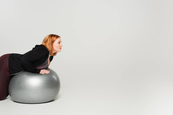 Vue latérale de l'entraînement femme taille plus sur balle de fitness sur fond gris - Photo, image
