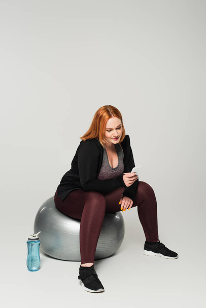 Body positive sportswoman using smartphone on fitness ball near sports bottle on grey background - Photo, Image