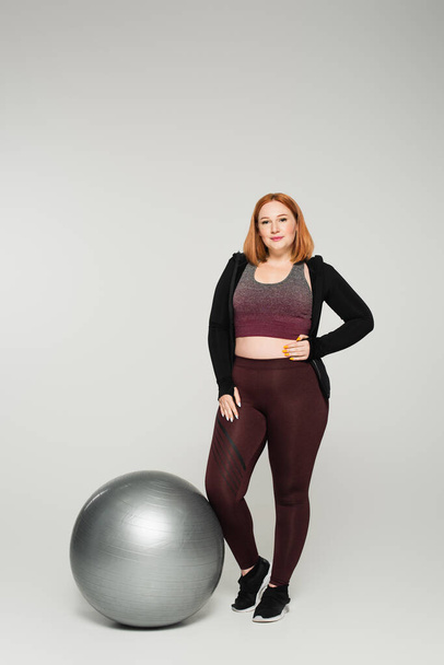 Plus size sportswoman holding hand on hip near fitness ball on grey background - Фото, изображение