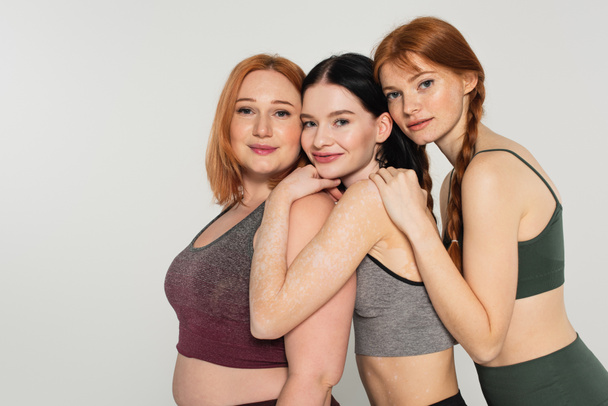 Young body positive friends in sportswear hugging isolated on grey  - 写真・画像