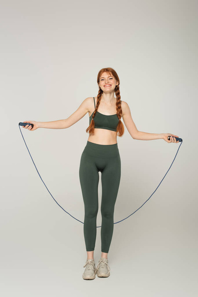 Smiling freckled sportswoman holding jump rope on grey background - Foto, immagini