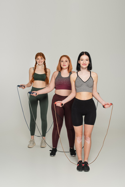 Happy body positive sportswomen holding jump ropes on grey background - Foto, imagen