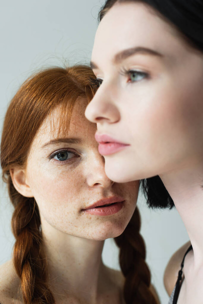 Young woman with freckles looking at camera near blurred friend isolated on grey  - Photo, Image
