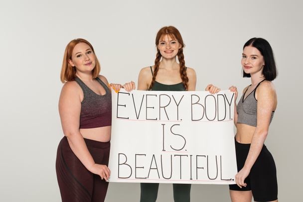 Usmívající se tělo pozitivní sportovkyněženy drží plakát s každým tělem v krásné písmo izolované na šedé  - Fotografie, Obrázek