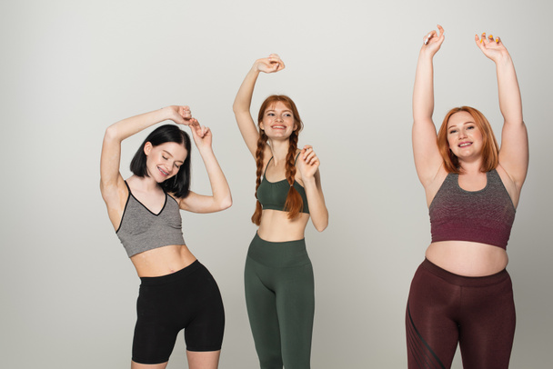 Fröhliche körperlich positive Sportlerinnen tanzen vereinzelt auf grau  - Foto, Bild