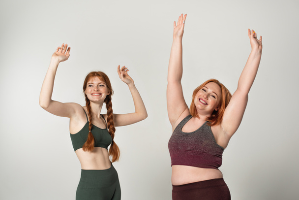 Smiling body positive sportswomen raising hands isolated on grey  - Foto, Imagem