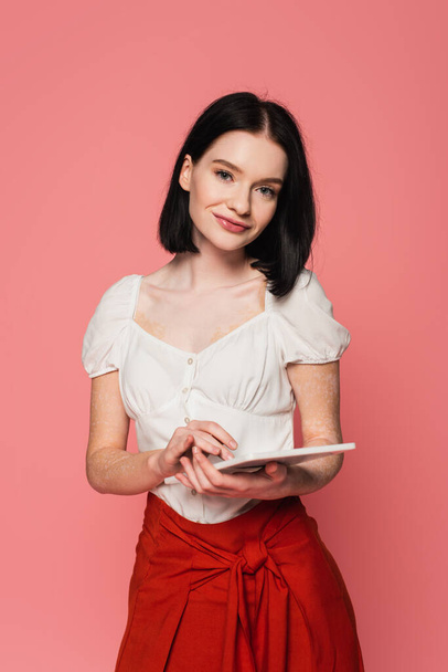 Smiling woman with vitiligo holding digital tablet isolated on pink  - Foto, afbeelding