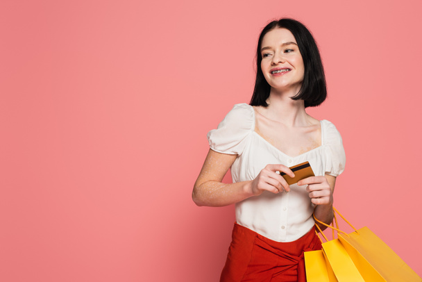 Šťastná žena s vitiligo držení kreditní karty a nákupní tašky izolované na růžové  - Fotografie, Obrázek