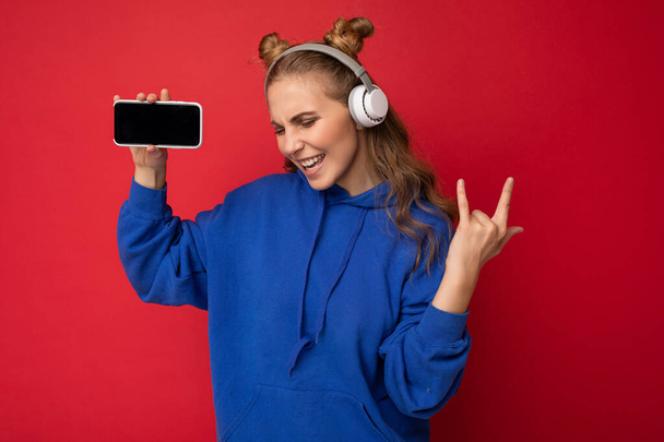 Приваблива позитивна усміхнена молода жінка в стильному повсякденному вбранні ізольована на барвистій стіні, що тримає і показує мобільний телефон з порожнім екраном для вирізання в білому Bluetooth
 - Фото, зображення